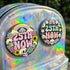 A round pinback button featuring a solid black background covered in a retro pastel colored design. The center reads 25TH NOW in a groovy bubbly font using pale pink and mint green referencing the 25th amendment. Surrounding the text is a maximalist cluster of cute but edgy motifs including smiling white skulls, pastel rainbows, pale yellow crescent moons, daisies, and a pink and white spotted mushroom. The button has a glossy laminate finish and a secure metal safety pin backing.