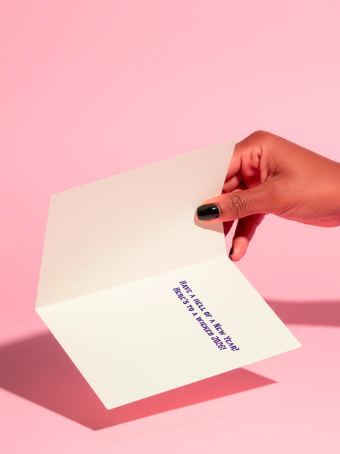 A vertical 5x7 inch folded greeting card featuring a detailed, psychedelic illustration of the folklore figure Krampus for a New Year celebration. The card stock has a shimmering pearlescent finish that reflects light, creating a glowing effect on the purple and teal artwork. The design mimics a vintage event poster reading "Krampus Ball 2026" and "Entry Fee: One Naughty Soul" in retro fonts. Krampus is depicted with large horns, sunglasses, and a long tongue holding a candy cane.