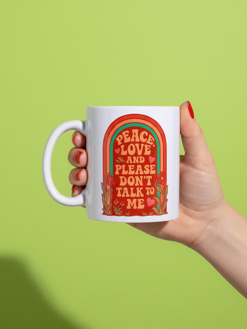 A glossy white porcelain ceramic mug featuring a vibrant maximalist design centered on the exterior. The artwork displays a retro 1970s-inspired rainbow arch composed of concentric bands in burnt orange, mustard yellow, and teal green. Inside the arch, stylized vintage bubble-serif typography in reddish-orange reads PEACE LOVE AND PLEASE DON'T TALK TO ME. The design evokes a groovy, hippie aesthetic mixed with sassy introvert humor.