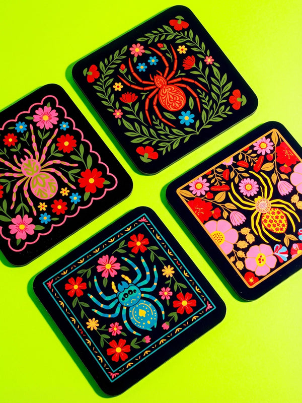 Set of four square coasters featuring a colorful folk art style design on a black background. Each coaster depicts a stylized spider in the center, surrounded by a symmetrical border of vibrant flowers and leaves. The spiders and florals are rendered in bright jewel tones of pink, blue, yellow, and red. The style mimics traditional tole painting with a maximalist, entomological twist. Shown on a flat surface to highlight the glossy finish and slim 0.125 inch profile.