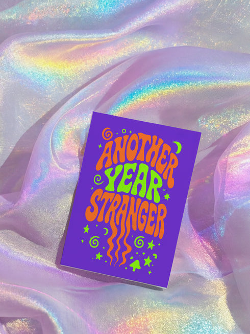 a purple book with the title "Another Year Stranger" printed on it, placed on a shimmering fabric surface.