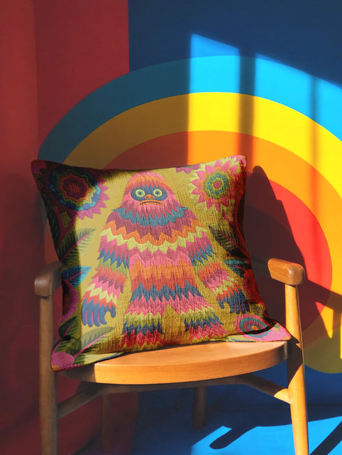A square faux embroidery pillow featuring a brightly colored maximalist design against a mustard yellow background. In the center stands a stylized, furry Bigfoot or Sasquatch figure rendered in vibrant layers of pink, orange, blue, and lime green with a blue face and yellow eyes. The central creature is surrounded by large, detailed symmetrical pink and blue flowers and leaves. The flat printed fabric creates a highly realistic optical illusion of dimensional crewel embroidery stitches.