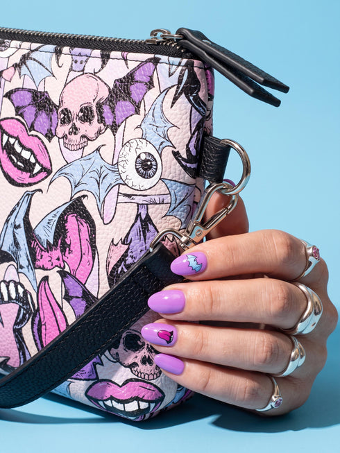 Small vegan leather crossbody purse with a real pebble grain texture and adjustable black strap. The flat printed artwork features a scattered all over pattern of pink skulls, purple and pastel blue winged bats, pink fanged mouths, and loose eyeballs set against a cream colored background. The bag is shown with black and silver hardware including a black top zipper, black zipper pull, black strap tabs, and silver metal rings and clips. The compact rectangular bag measures 8.5 by 7 by 2 inches.