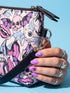 Small vegan leather crossbody purse with a real pebble grain texture and adjustable black strap. The flat printed artwork features a scattered all over pattern of pink skulls, purple and pastel blue winged bats, pink fanged mouths, and loose eyeballs set against a cream colored background. The bag is shown with black and silver hardware including a black top zipper, black zipper pull, black strap tabs, and silver metal rings and clips. The compact rectangular bag measures 8.5 by 7 by 2 inches.