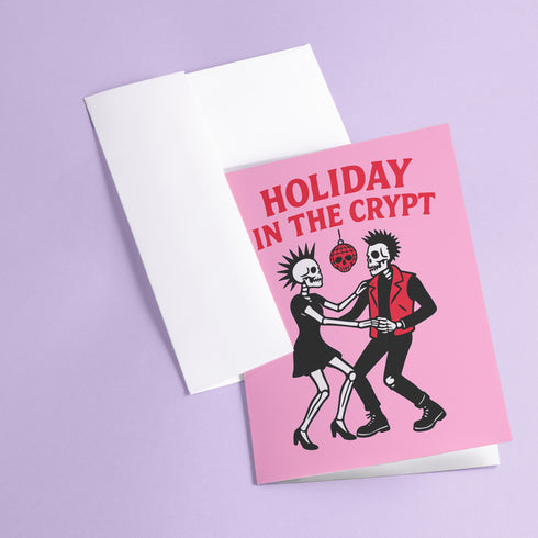 A 5x7 inch greeting card with a vibrant pink background and red retro text reading 'HOLIDAY IN THE CRYPT'. The illustration shows a male and female punk rock skeleton dancing under a red skull disco ball. The female skeleton has a mohawk and wears a black dress. The male skeleton has a mohawk and wears a black jacket with a red vest. Inside, the message reads, 'From our crypt to yours. Eat, drink, and be scary.' The style is a graphic, punk aesthetic.