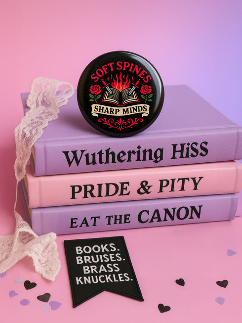 A round pinback button in black with a glossy mylar face and metal construction. The design, in a traditional tattoo style, features a central open book with flames rising from its pages. Two silver swords with black hilts are crossed over the book. On either side of the book is a red rose with a green stem and leaves. Below the book is a cream-colored banner with the words "SHARP MINDS" in a black serif font. Above the book, the words "SOFT SPINES" are written in a curved, red font.