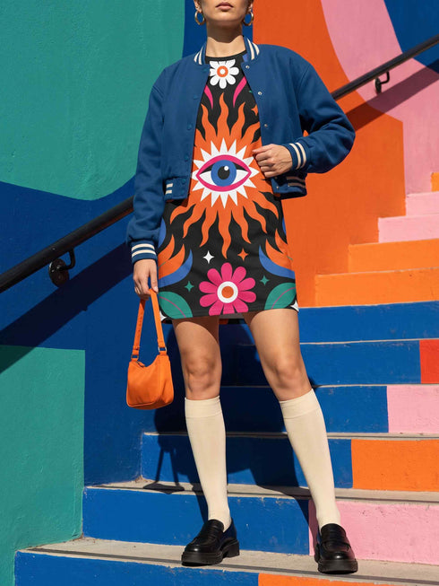 A short-sleeve, above-the-knee t-shirt dress with a straight, structured fit. The dress has a true black background featuring a bold, vibrant graphic pattern. The center displays a large blue and white evil eye surrounded by a bright orange wavy sunburst with a white jagged border. Above and below the eye are bright pink daisy flowers with orange centers, scattered white four-petal stars, and wavy blue and teal green leaf shapes. The fabric is thick, smooth, and holds its clean shape.