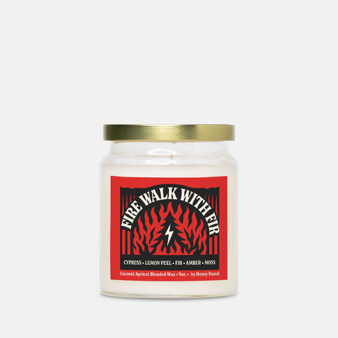 A 9-ounce hand-poured candle inspired by the TV show Twin Peaks, named "Fire Walk With Fir." The candle is made from a creamy, off-white coconut apricot blended wax, housed in a clear glass vessel with a classic, slightly tapered shape. A prominent matte-finish label on the front features the title in a stylized font over a red and black graphic with flame and fir tree motifs. The label also lists scent notes of cypress, lemon peel, fir, amber, and moss.