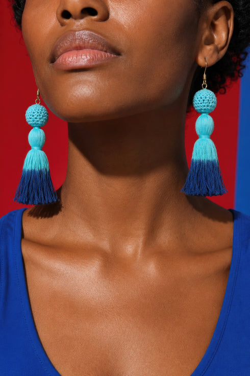 A close-up of a woman's face wearing large, blue tassel earrings with a blue tassel at the end.