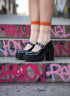 A pair of black high-heeled shoes with a chunky sole, worn with pink and yellow checkered socks, standing on a concrete step with graffiti on it.