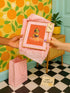 A vibrant fine art print of a rubber duck inspired by Frida Kahlo, complete with bold unibrow, floral crown, and traditional embroidered blouse. Framed in a red sculptural frame, the duck floats in orange-tinted bubbles against a deep orange backdrop and violet wall.