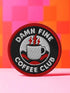 A round, 3-inch embroidered patch with a thick, red merrowed border for a clean, finished edge. The design features a solid black background with the words 'DAMN FINE' in an arc at the top and 'COFFEE CLUB' in an arc at the bottom, all in a bold, white, sans-serif font. In the center, a detailed white coffee mug with a black handle is depicted. Two bright red lightning bolts emerge from the top of the mug, symbolizing energy, reminiscent of the Black Lodge floor from Twin Peaks.