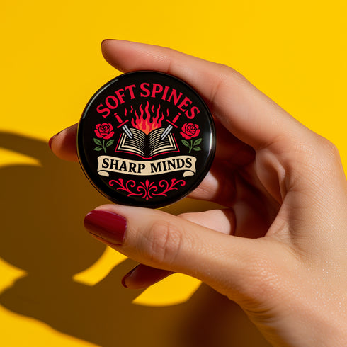 A round pinback button in black with a glossy mylar face and metal construction. The design, in a traditional tattoo style, features a central open book with flames rising from its pages. Two silver swords with black hilts are crossed over the book. On either side of the book is a red rose with a green stem and leaves. Below the book is a cream-colored banner with the words "SHARP MINDS" in a black serif font. Above the book, the words "SOFT SPINES" are written in a curved, red font.