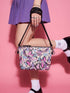 Small vegan leather crossbody purse with a real pebble grain texture and adjustable black strap. The flat printed artwork features a scattered all over pattern of pink skulls, purple and pastel blue winged bats, pink fanged mouths, and loose eyeballs set against a cream colored background. The bag is shown with black and silver hardware including a black top zipper, black zipper pull, black strap tabs, and silver metal rings and clips. The compact rectangular bag measures 8.5 by 7 by 2 inches.