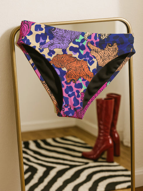 Psychedelic bikini bottom with multicolored tiger print on a neon leopard background; mid-rise cut with black lining and vibrant color blocking in purple, blue, pink, orange, and cream; made of recycled polyester and spandex blend with cheeky fit.