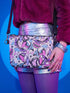 Small vegan leather crossbody purse with a real pebble grain texture and adjustable black strap. The flat printed artwork features a scattered all over pattern of pink skulls, purple and pastel blue winged bats, pink fanged mouths, and loose eyeballs set against a cream colored background. The bag is shown with black and silver hardware including a black top zipper, black zipper pull, black strap tabs, and silver metal rings and clips. The compact rectangular bag measures 8.5 by 7 by 2 inches.