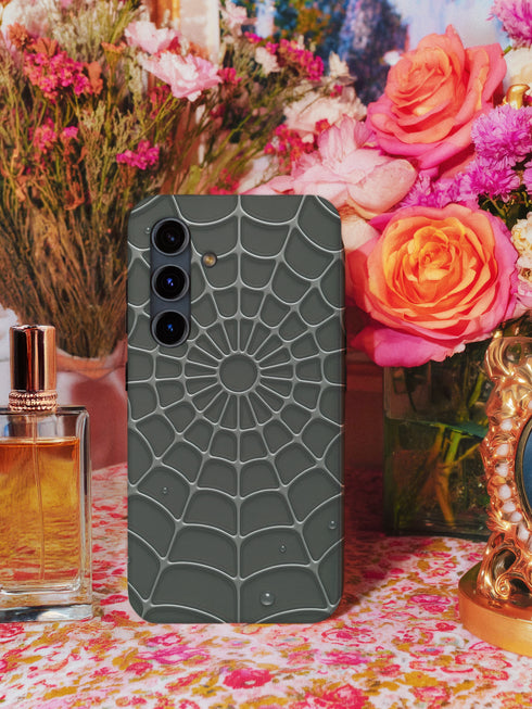 a smartphone case with a spider web design, placed on a table with a vase of flowers and a bottle of perfume.