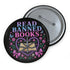 Glossy black pinback button that says “Read Banned Books” in purple and teal letters, featuring a chained book, roses, bows, and floral accents, styled on colorful books and pastel backgrounds.