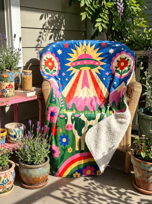 A rectangular velveteen minky blanket with a bright, maximalist design. The top half features a vibrant blue sky with a pink, green, and yellow UFO beaming rays down over hot pink snow-capped mountains. The bottom half shows a grassy green field with a white cow covered in dark spots, surrounded by large, colorful red, pink, and blue retro flowers, pine trees, and a rainbow arch at the very bottom. The reverse side is a fluffy off-white sherpa.