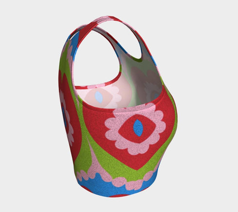 A sleeveless scoop neck crop top featuring a bold all-over printed design. The central graphic is a large bright red heart enclosing a scalloped pink flower shape and a blue evil eye motif in the middle. The background surrounds the heart with mirrored concentric wavy arches and scalloped borders in vibrant shades of lime green, pink, blue, and red. The fitted garment has a smooth athletic texture. Available in a standard matte performance knit or a sparkly silver glitter foil finish.