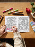 A 8.5 by 11 inch softcover coloring book titled The Feminist Rage Coloring Book For Cute Loud Unhinged Women. The black cover features vibrant pink and green typography, a fanged heart engulfed in flames, pink roses, and crescent moons. The back cover displays a heart pierced by a dagger and text reading Your feelings are valid your rage is too. Inside pages contain black and white line art with bold feminist quotes, floral borders, potions, coffins, and edgy witchy illustrations.