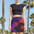 A woman wearing a black crop top and a colorful, patterned skirt stands in front of palm trees.