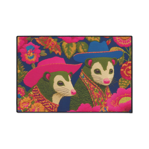 A rectangular non-slip doormat with a black trimmed edge. The mat features a vibrant, colorful folk art illustration on a dark navy blue background with a faux embroidered texture. The design depicts two friendly opossums, rendered in an olive green color with white faces and pink noses. They are surrounded by large, stylized flowers and leaves in shades of hot pink, red, turquoise, and yellow, creating a busy, maximalist pattern. The overall aesthetic is eclectic, whimsical, & highly detailed.