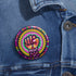 Round pinback button with psychedelic rainbow design featuring a raised black fist surrounded by concentric circles in vibrant pink, orange, green, and yellow. The word "RESIST" appears in white text below the fist, set against black background with flower details. Activist protest pin with glossy finish and safety pin backing.