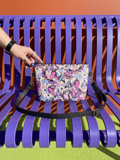 Small vegan leather crossbody purse with a real pebble grain texture and adjustable black strap. The flat printed artwork features a scattered all over pattern of pink skulls, purple and pastel blue winged bats, pink fanged mouths, and loose eyeballs set against a cream colored background. The bag is shown with black and silver hardware including a black top zipper, black zipper pull, black strap tabs, and silver metal rings and clips. The compact rectangular bag measures 8.5 by 7 by 2 inches.