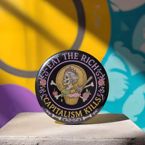Glossy black pinback button featuring a pastel embroidered-style design of a skeleton queen on a cupcake, surrounded by roses and bones, with the text “Eat the Rich” and “Capitalism Kills.” Displayed on protest props, tea cups, and pastel backgrounds.