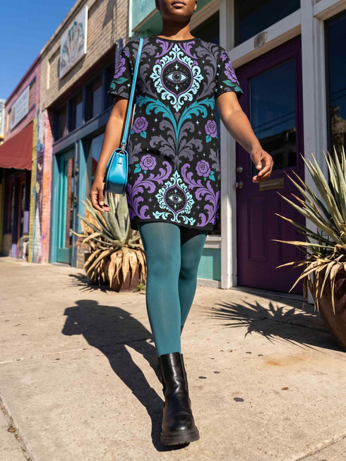 A short-sleeve structured t-shirt dress with a black background. The dress features a bold, vertically stacked repeating graphic print. The design includes intricate damask patterns in teal and grey, purple roses with teal leaves, and a central evil eye motif set inside an ornate frame with small four-point stars. The heavyweight fabric holds a clean, straight cut above the knee with zero drape, maintaining a sculptural, slightly boxy silhouette.
