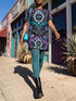 A short-sleeve structured t-shirt dress with a black background. The dress features a bold, vertically stacked repeating graphic print. The design includes intricate damask patterns in teal and grey, purple roses with teal leaves, and a central evil eye motif set inside an ornate frame with small four-point stars. The heavyweight fabric holds a clean, straight cut above the knee with zero drape, maintaining a sculptural, slightly boxy silhouette.