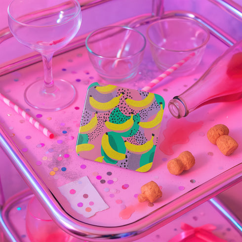 Square drink coaster featuring a playful banana print with bold yellow bananas over a pastel pink, purple, and green abstract background. Made of durable hardboard MDF with a cork backing and a glossy protective topcoat. Measures 3.74 by 3.74 inches, water-repellent and heat-resistant. Perfect for fruit-themed, retro, Y2K, coquette, or dopamine-core decor styles.