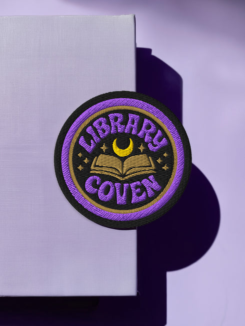3-inch round embroidered iron-on patch in a cotton–polyester blend; black base with purple ring and gold accent ring; open book, gold crescent moon, tiny stars; curved purple text reads “LIBRARY COVEN”; merrowed edge. Witchy book lover patch for librarians, dark academia, and BookTok covens; makes a great gift for readers, witches, and spooky library pals.
