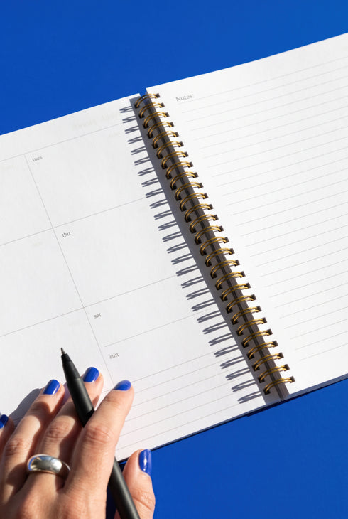 A hand holding a black pen is seen writing in a spiral-bound notebook with a blue background.