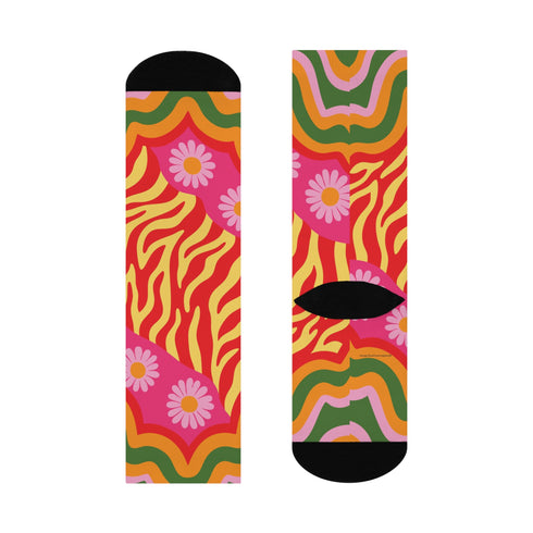 A pair of vibrant crew socks featuring a maximalist 1970s inspired collage pattern. The design includes blocks of hot pink with white daisy flowers and yellow centers, clashing playfully with panels of mustard yellow and orange tiger stripes. Wavy psychedelic lines in grass green and orange frame the top cuff. The socks have a solid black heel and toe cap. The fabric is a smooth, stretchy polyester blend with a flat knit texture and bright colors.