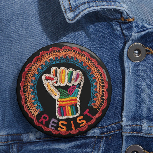 Round pinback metal button with an embroidered-style design on a black background. The center features a raised fist symbol stitched in a multicolored pattern, incorporating rainbow hues and folk-inspired textures. The fist is encircled by a vibrant, lace-like border in shades of blue, orange, and red, resembling intricate embroidery. Below the fist, the word "RESIST" is spelled out in bold, stitched-textured letters in red and orange. The button has a silver metal back with a secure pin closure.
