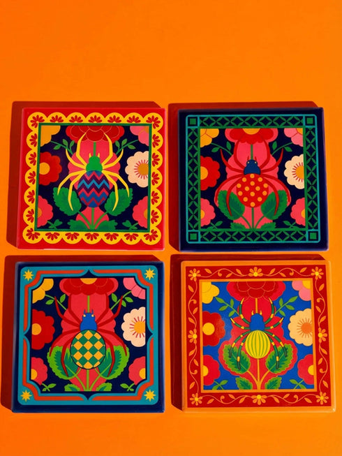 A set of four square coasters featuring a glossy glazed finish and vibrant insect illustrations. Each coaster measures approximately four by four inches. The designs showcase symmetrical beetles and spiders in bold red, blue, and orange colors, framed by intricate floral and geometric borders in a maximalist folk art style. Available in ceramic with cork backing or hardboard MDF. The edges are smooth. The designs are vivid and highly saturated, designed to provide grip and protect furniture.