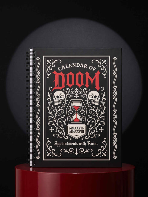 An 8.5 x 11 inch black hardcover spiral planner with a durable satin matte laminated finish. The front cover features a vintage-style white ornamental border, creating a gothic, macabre aesthetic. Inside the frame, the title "Calendar of Doom" is printed in an antique serif font. This undated weekly and monthly agenda is shown with a metal wire-o spiral binding on the right side. The design uses a black, white, and red color scheme, making it a unique piece of dark humor stationery