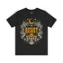 A unisex short sleeve graphic t shirt in dark charcoal black featuring a highly detailed symmetrical medallion print on the chest. In the center, the word RESIST is printed in a bold yellow gothic blackletter font. Two coiled white snakes with fine black scale details frame the text on the left and right. Above the text, a yellow crescent moon is flanked by white and yellow starbursts. Beneath, three yellow marigold flowers sit atop white stems and leafy foliage.