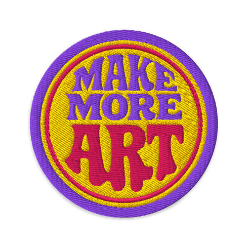 A round embroidered patch with the words "MAKE MORE ART" in a retro 70s psychedelic font. The word "ART" is magenta pink, "MAKE MORE" is purple, and they are set against a bright yellow circular background. The entire design is encircled by a thin pink line and a thick purple border. This 3-inch iron-on applique is a colorful, groovy accessory for DIY projects, perfect for personalizing jackets, backpacks, or denim vests. It serves as an inspirational artist gift with a vintage aesthetic.