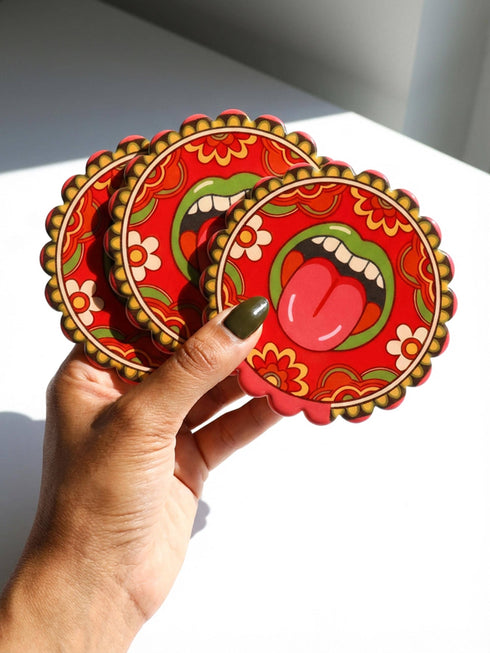 Round ceramic coaster with scalloped edges featuring a vibrant pop art design of an open mouth with green lips and a red tongue sticking out. The background is a bright red with psychedelic floral swirls in yellow orange and green. The surface is glossy glazed ceramic. The back is made of cork to prevent slipping. This maximalist decor piece fits retro 70s funky and weirdcore aesthetics. It is a bold statement piece for coffee tables. The design is colorful and eclectic dopamine decor.