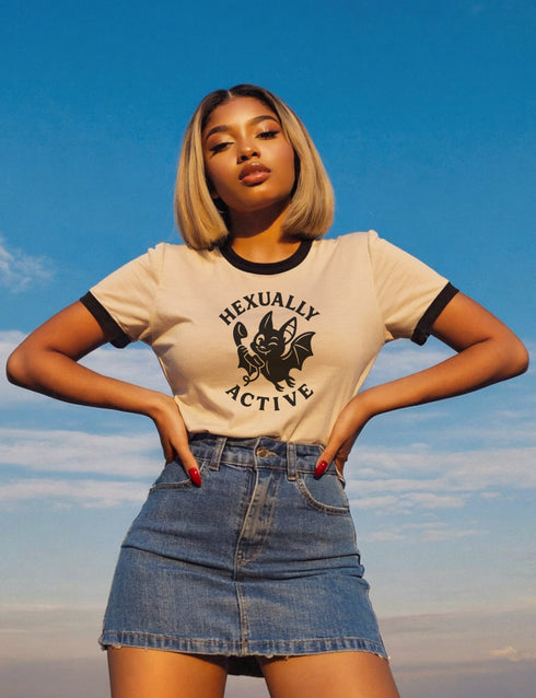 Unisex offwhite and black ringer t-shirt with a bold black graphic of a cartoon bat holding a wand, surrounded by the phrase “Hexually Active.” The design is playful, witchy, and gothic, perfect for lovers of magic, spellwork, hexes, and witch humor. The shirt features contrast collar and sleeves in black for a retro vibe.