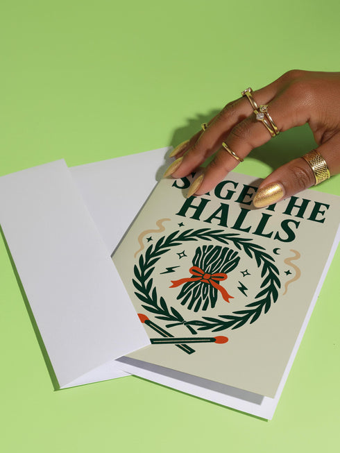 A detailed folk-art style illustration of a "Sage the Halls" holiday greeting card. The design features a bundle of sage smudge sticks tied with a red ribbon at the center of a dark green wreath. The wreath is accented with small crescent moons, stars, and smoke swirls. Below the wreath are two crossed matches with red tips. The card is printed on creamy beige paper with bold, dark green serif text. This funny spiritual and witchy Yule card is a unique pagan greeting for energy cleansing.
