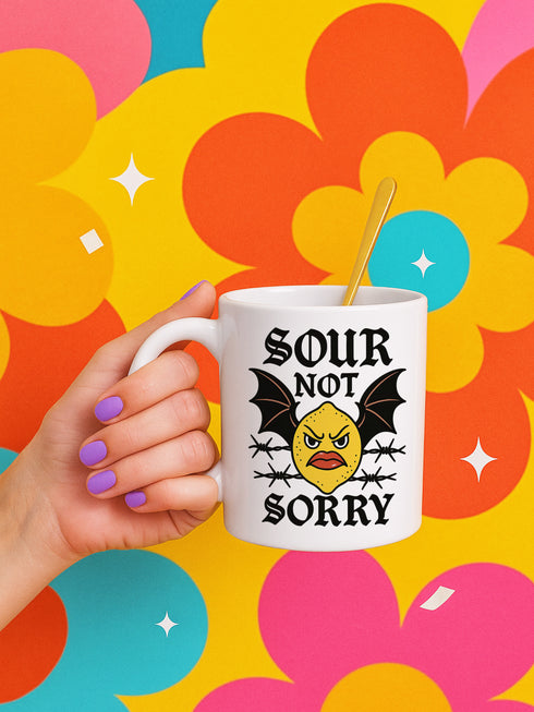 White ceramic 11oz coffee mug featuring a bold tattoo-style graphic with an angry yellow lemon face, black bat wings, and barbed wire details. The design reads “SOUR NOT SORRY” in gothic black lettering above and below the lemon. C-handle, durable construction, dishwasher and microwave safe. Graphic is high-contrast and punk-inspired, perfect for adding attitude to your drinkware collection.
