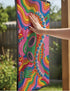 A rectangular doormat featuring a vibrant 2D flat print designed to look like textured yarn embroidery. The central graphic showcases a bright green alien with large black eyes, wearing a pink neck bandana and a highly decorated red, pink, and blue sombrero cowboy hat. The alien has one green hand raised in a friendly wave. The background is a psychedelic pattern of wavy, flowing rainbow stripes in yellow, orange, red, pink, blue, and green. The mat has a sleek black outer edge trim.