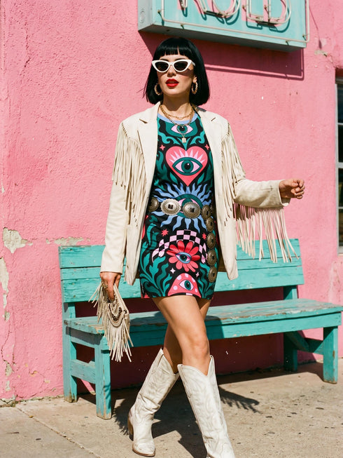 A short-sleeve, above-the-knee t-shirt dress with a true black background. The front features a vibrant, vertically stacked symmetrical graphic design. From top to bottom, the design includes a pink heart containing a green and blue eye, a large blue sunburst with a central eye, a red daisy flower with an eye flanked by pink and black checkered arches, and a pink mushroom cap with an eye. Swirling emerald green foliage, pink stars, and purple stars fill the negative space around the motifs.