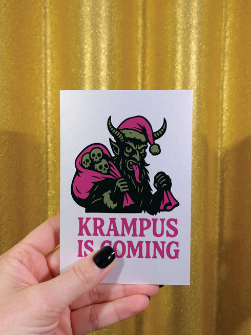 A 5x7 inch Krampus Christmas card shown in two paper finishes. The front features a stylized illustration of the folklore demon Krampus with green skin, black horns, and a pink Santa hat. He carries a large pink sack filled with three skulls over his shoulder. Below him, the words "KRAMPUS IS COMING" are printed in bold pink text. The inside of the card is blank on the left and has the printed text "Hope you've been beautifully bad this year." on the right.