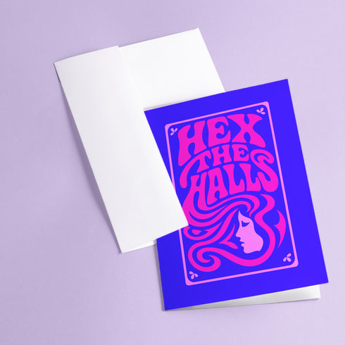 A vertical 5x7 greeting card made from premium 120lb silk cover stock with a slight sheen. The card has a deep purple background with a thin pink border. In the center, the words 'Hex the Halls' are written in a bubbly, psychedelic 70s-style font in shades of hot pink and light pink. Below the text is a stylized pink silhouette of a woman's face in profile with long, flowing, wavy hair. The design is reminiscent of retro witchcraft and groovy aesthetics, perfect for an alternative holiday.
