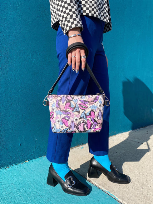 Small vegan leather crossbody purse with a real pebble grain texture and adjustable black strap. The flat printed artwork features a scattered all over pattern of pink skulls, purple and pastel blue winged bats, pink fanged mouths, and loose eyeballs set against a cream colored background. The bag is shown with black and silver hardware including a black top zipper, black zipper pull, black strap tabs, and silver metal rings and clips. The compact rectangular bag measures 8.5 by 7 by 2 inches.